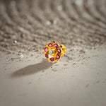 An 18K gold nose pin shaped like a small flower with ruby-red petals and a clear crystal center, photographed on a light textured surface