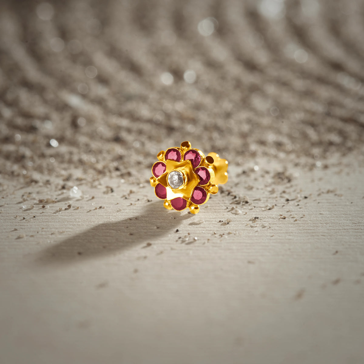 An 18K gold nose pin shaped like a small flower with ruby-red petals and a clear crystal center, photographed on a light textured surface