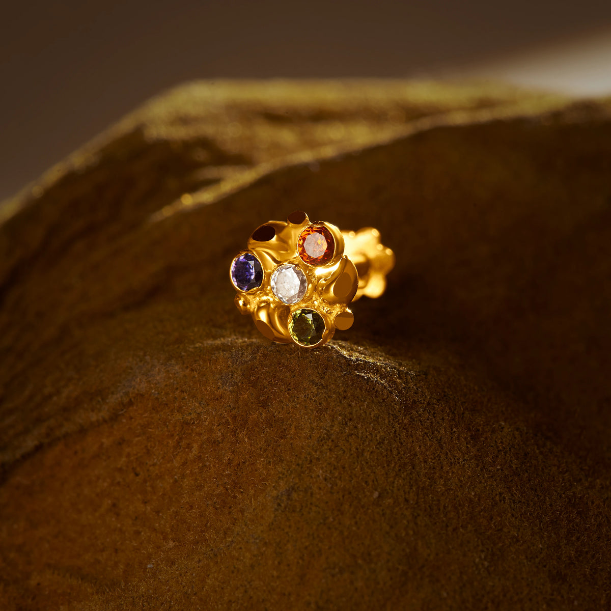 An 18K gold nose pin with a textured crown-like setting, set with multicolored stones in blue, white, orange, and green, photographed on a dark textured fabric background