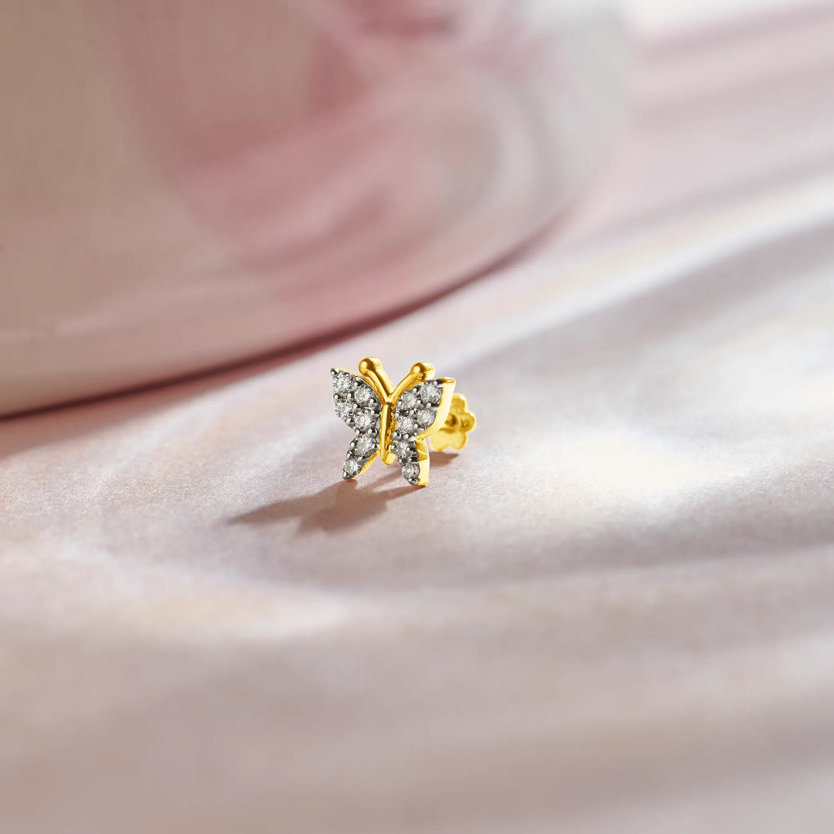 Design of gold nose pin with butterfly shape and diamond detailing on soft pink fabric background