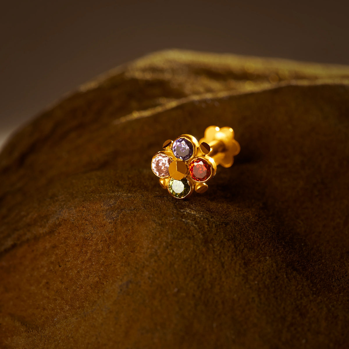 An 18K gold nose pin shaped like a small four-petal flower, set with multicolored stones in purple, pink, red, and green, photographed on a dark textured fabric background