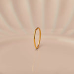 Front view of slim 18K gold hoop nose ring standing upright on a soft beige background with subtle light rays