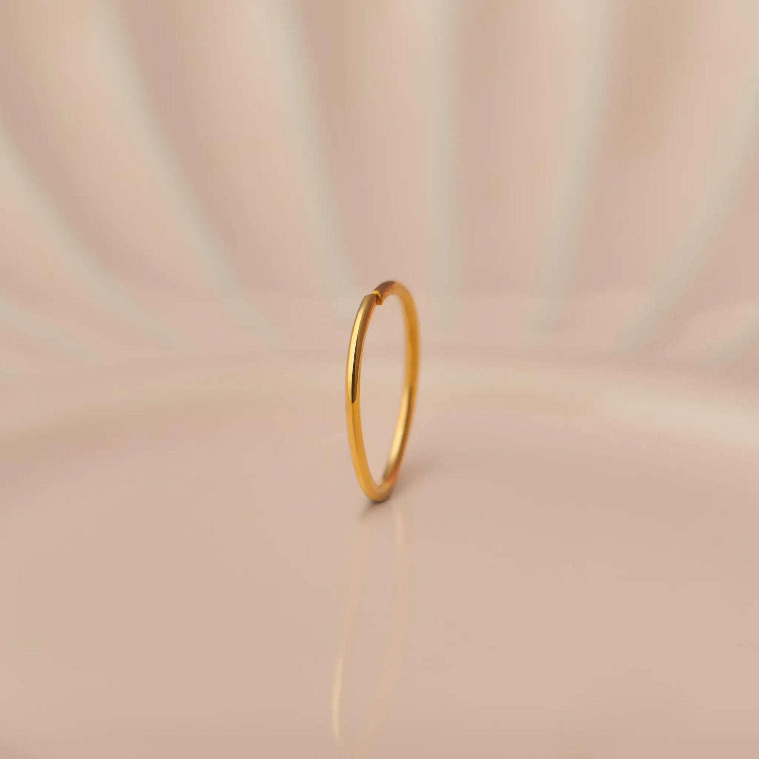 Front view of slim 18K gold hoop nose ring standing upright on a soft beige background with subtle light rays
