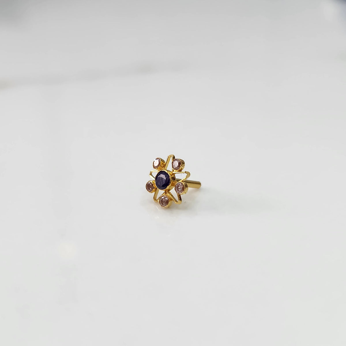 A gold nose pin shaped like a star with an oval deep purple center stone and light champagne accent stones