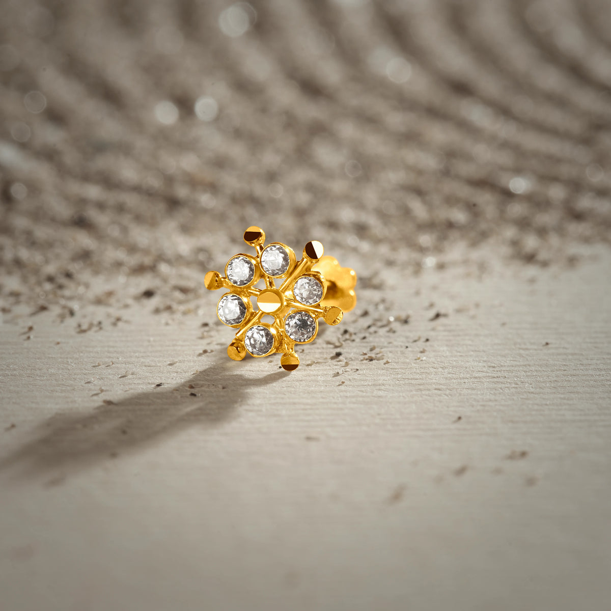 An 18K gold nose pin shaped like a wheel with pointed gold spokes between clear crystal stones, photographed on a light textured surface