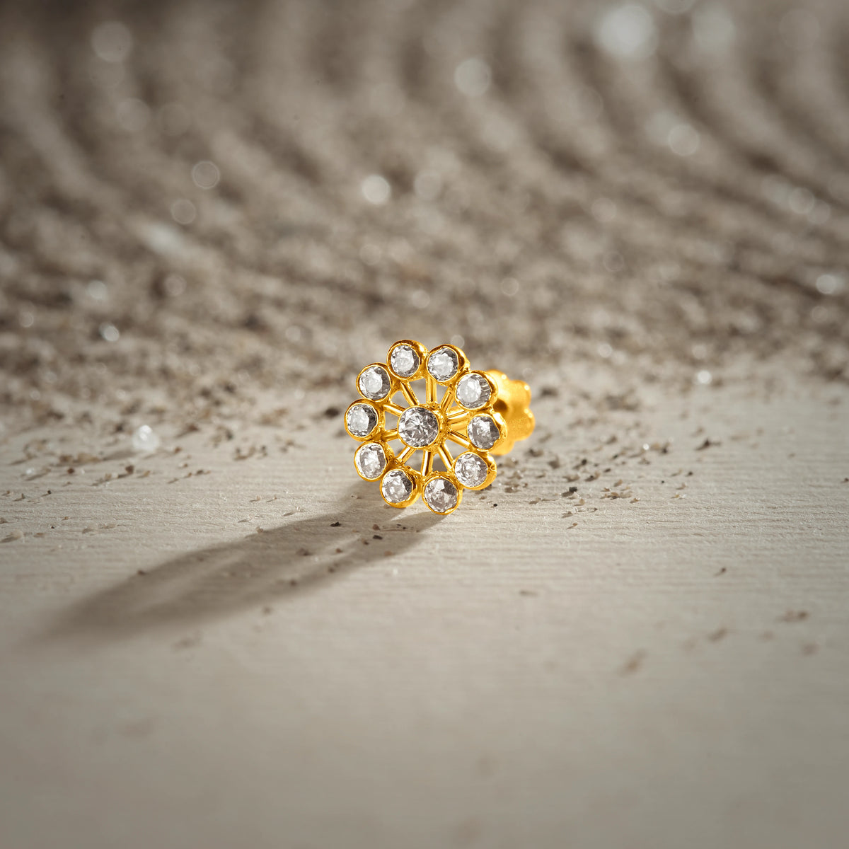 An 18K gold nose pin shaped like a spoked wheel with open petal loops, set with clear crystal stones, photographed on a light textured surface