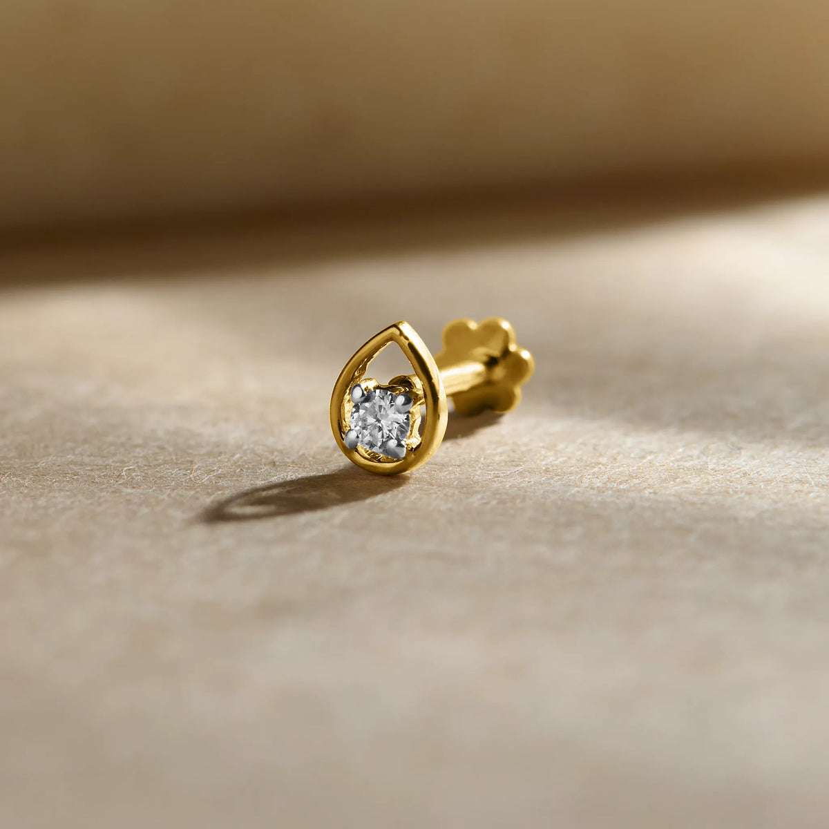 Gold nose pin design with elegant teardrop shape featuring a sparkling solitaire stone set in a polished gold base on a soft neutral background