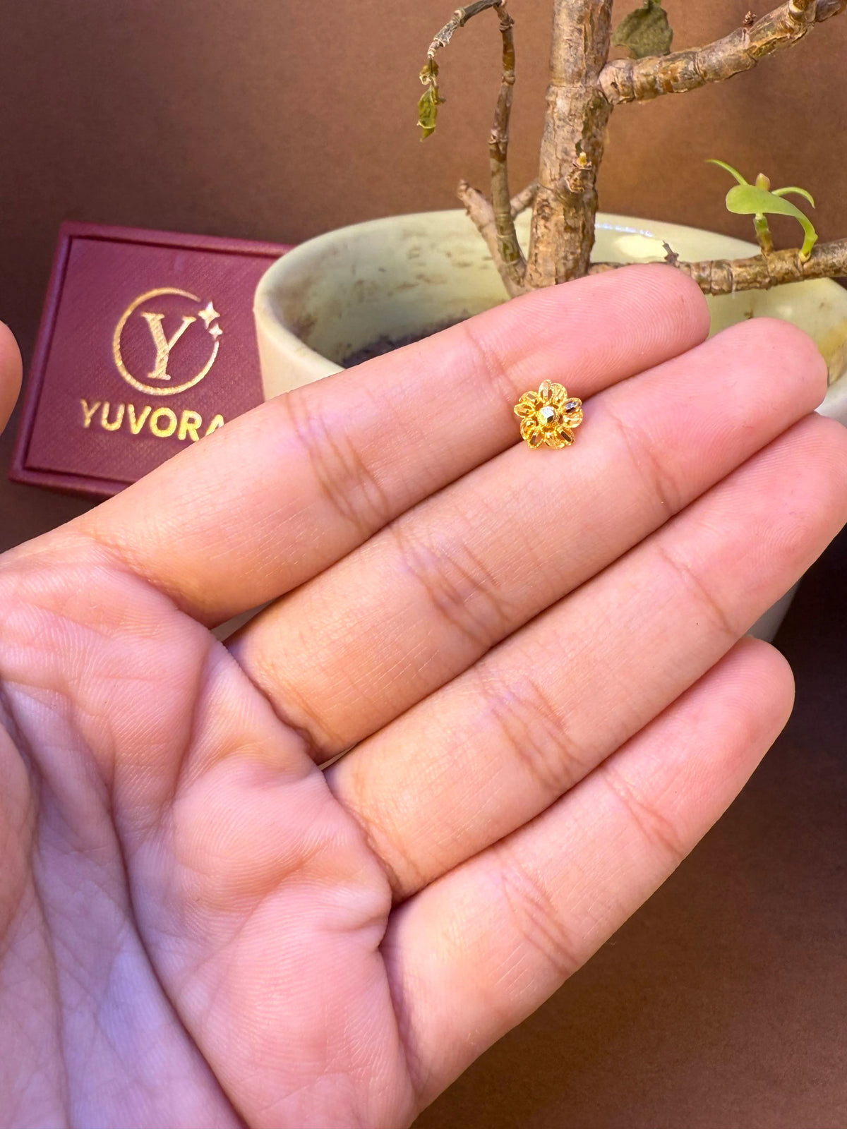 Round flower-shaped gold nose pin with center stone held in hand, Yuvora jewelry packaging in background