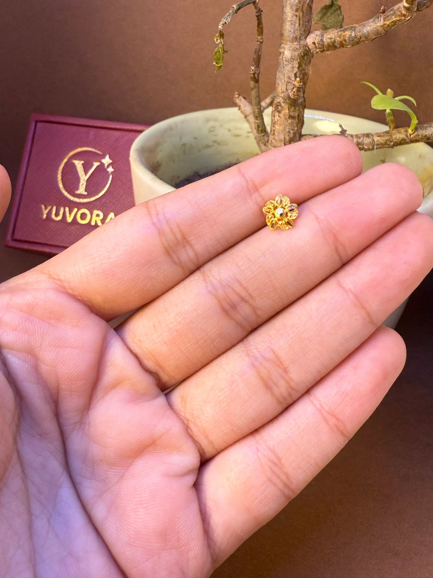 Round flower-shaped gold nose pin with center stone held in hand, Yuvora jewelry packaging in background
