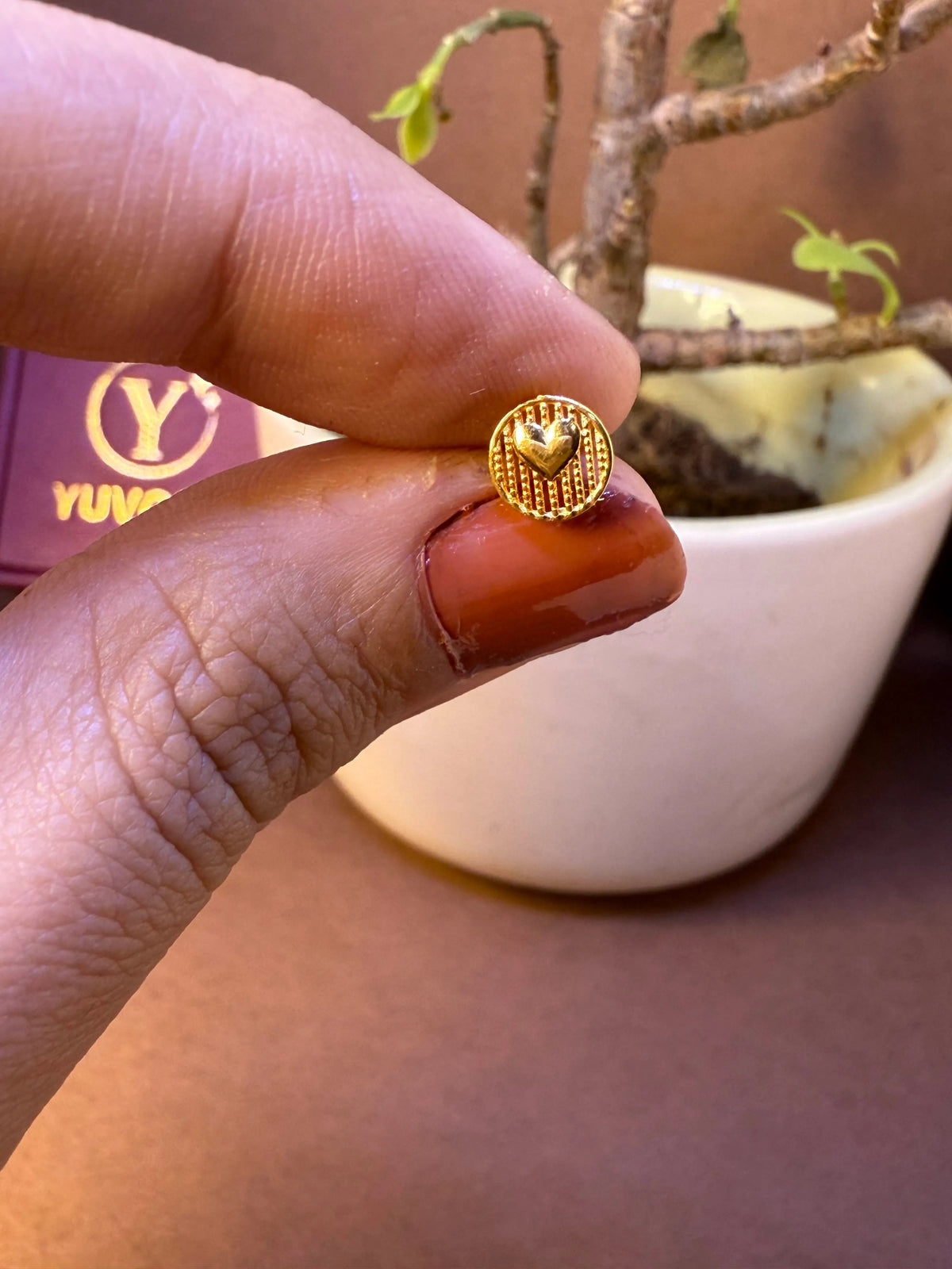 Round textured gold nose pin with raised heart center, held between fingertips