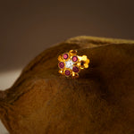 An 18K gold nose pin shaped like a flower with ruby-red petals and a clear crystal center, photographed on a dark textured fabric background