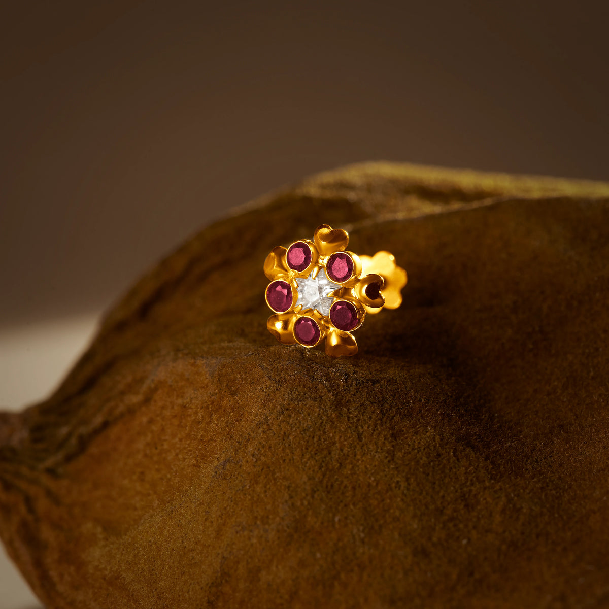 An 18K gold nose pin shaped like a flower with ruby-red petals and a clear crystal center, photographed on a dark textured fabric background