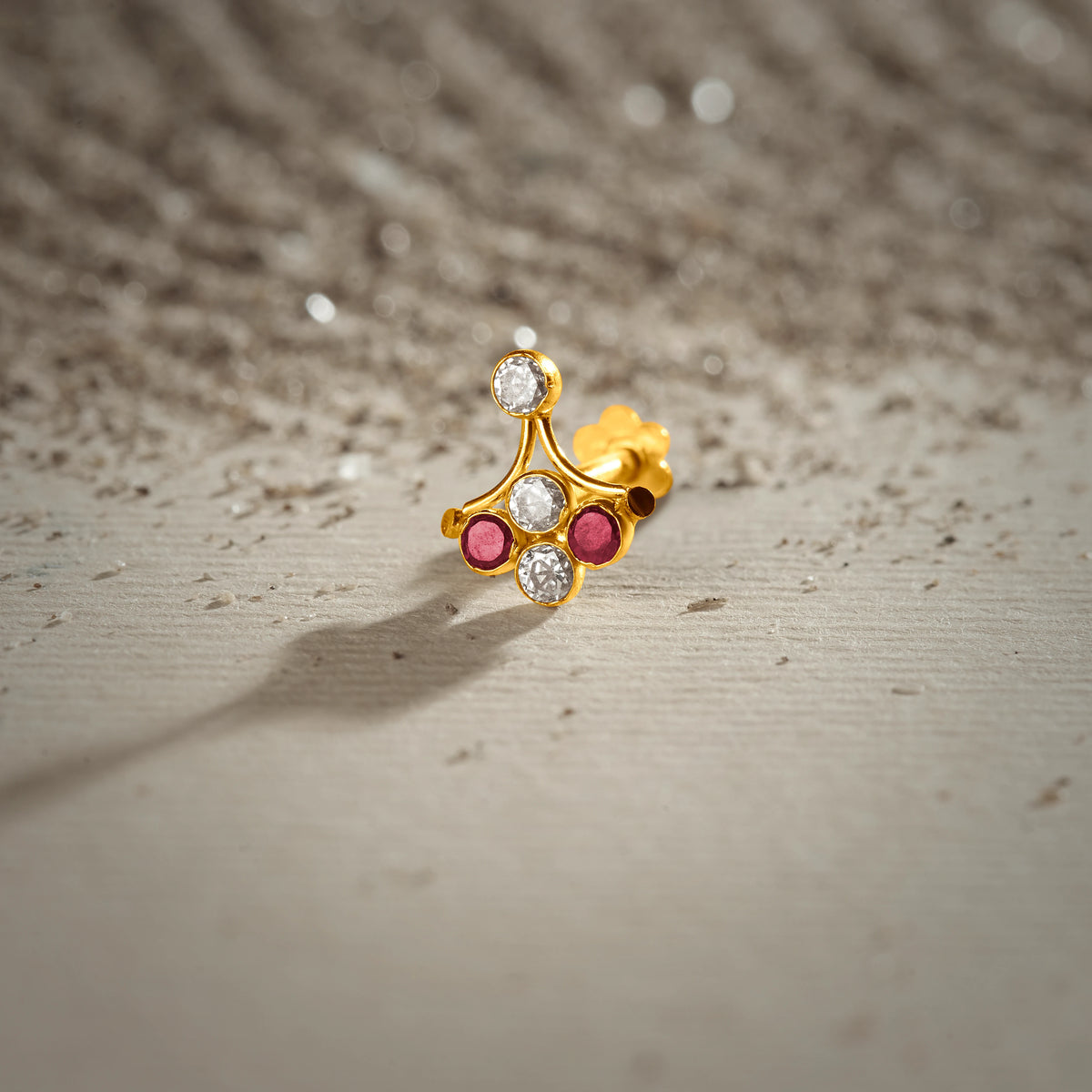 An 18K gold nose pin with a fan-shaped leaf design, set with alternating clear and ruby-red stones, photographed on a light textured surface