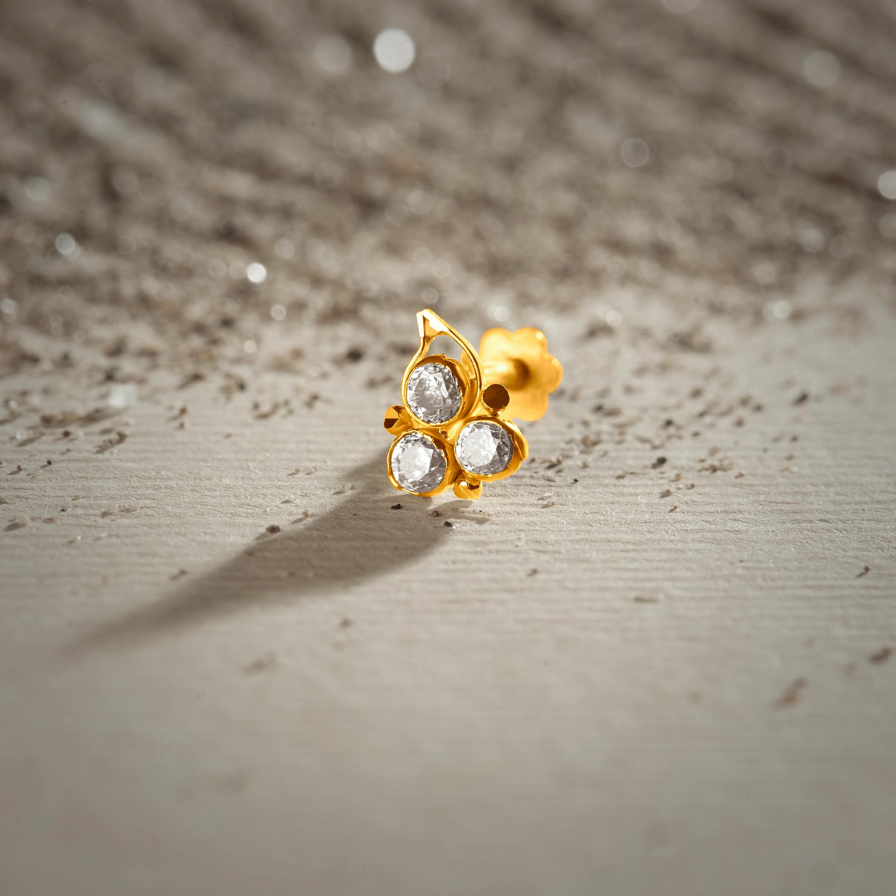 An 18K gold nose pin with a trefoil design featuring an open teardrop loop and three clear crystal stones, photographed on a light textured surface