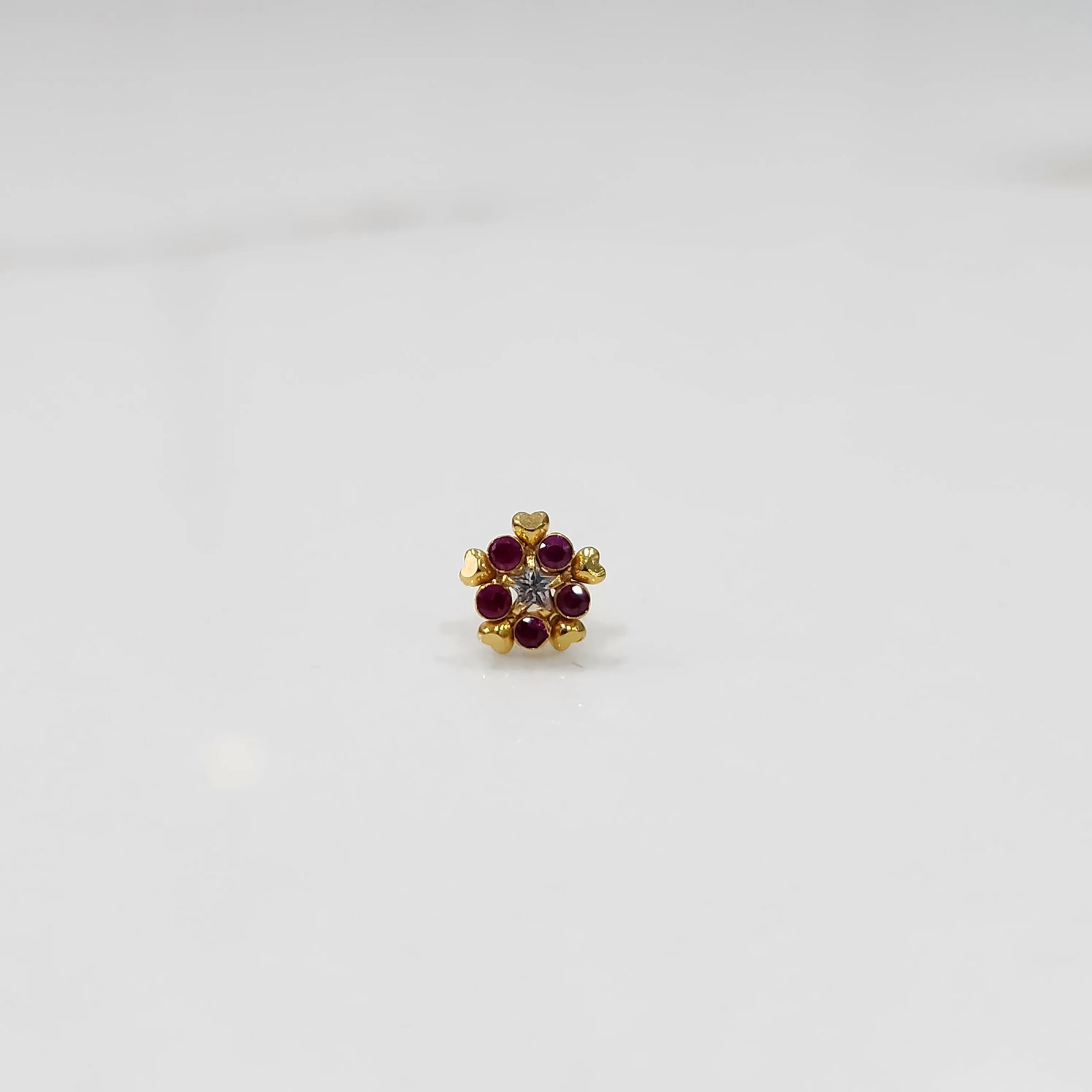 Product shot of a gold nose stud with ruby flower and heart petal design
