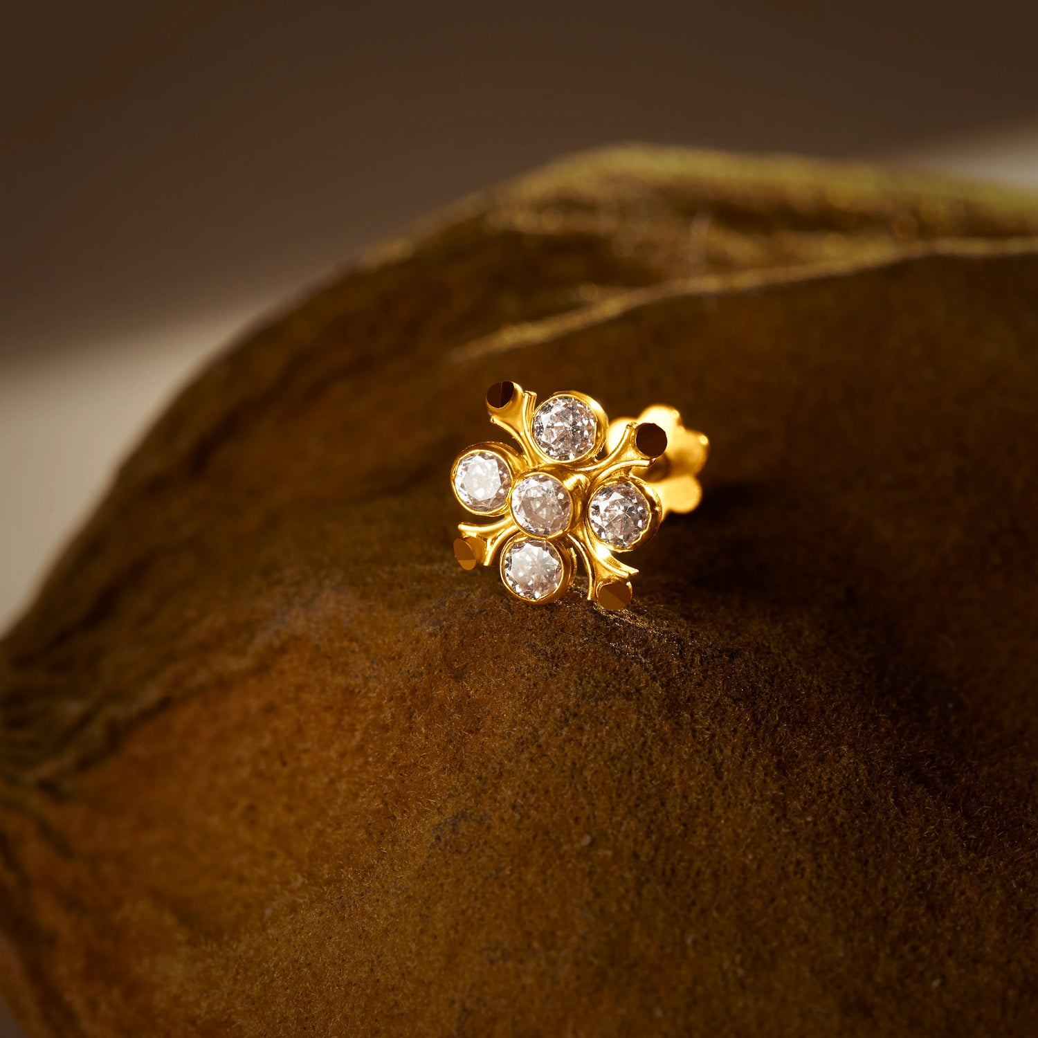 An 18K gold nose pin shaped like a flower cluster, set with clear crystal stones, photographed on a dark textured fabric background