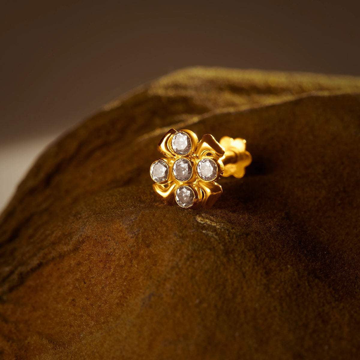 An 18K gold nose pin shaped like a flower with clear crystal petals, photographed on a dark textured fabric background