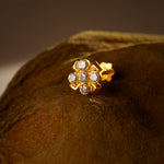 An 18K gold nose pin shaped like a flower with clear crystal petals, photographed on a dark textured fabric background