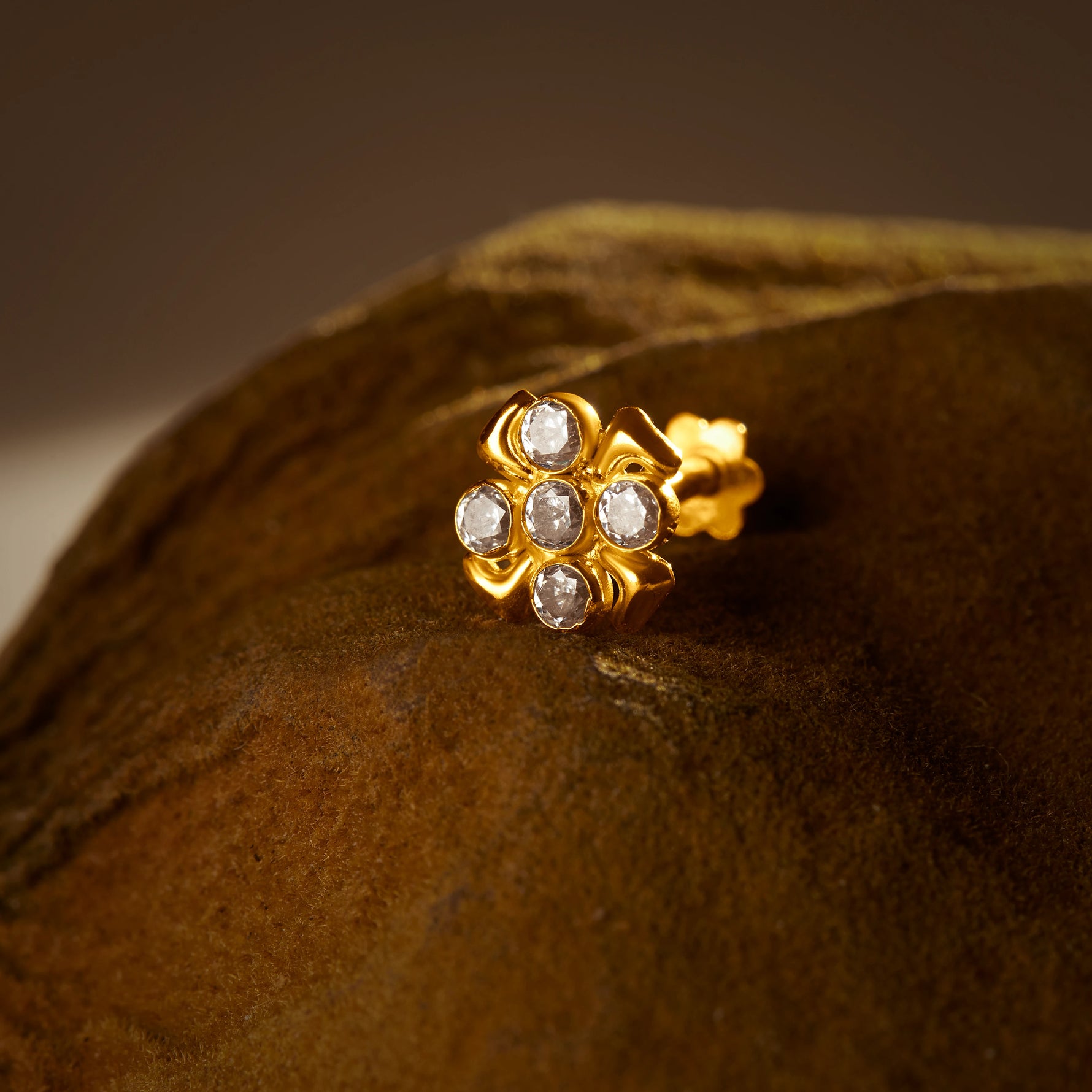 An 18K gold nose pin shaped like a flower with clear crystal petals, photographed on a dark textured fabric background