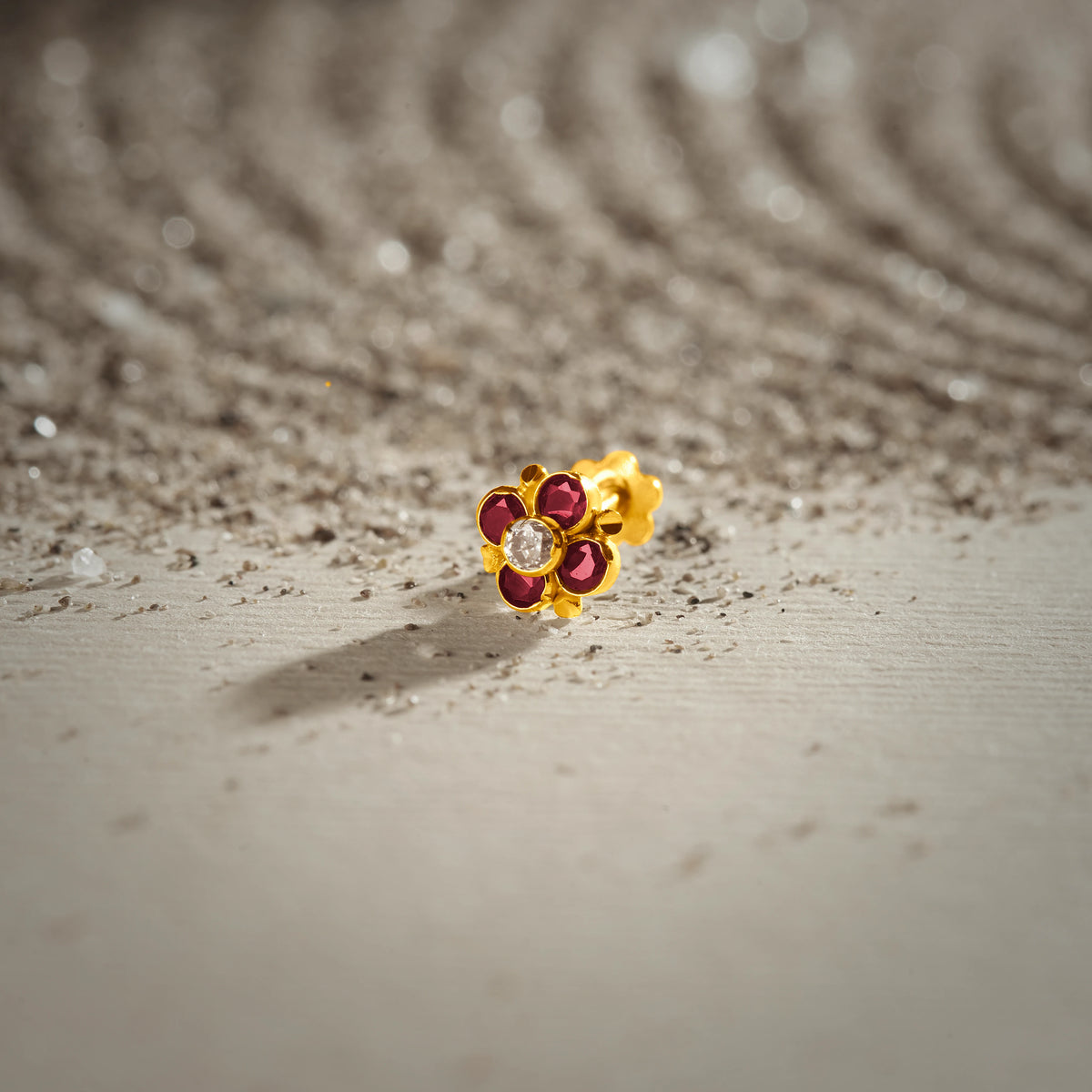 An 18K gold nose pin shaped like a flower with ruby-red petals and a clear crystal center, photographed on a sandy textured surface 