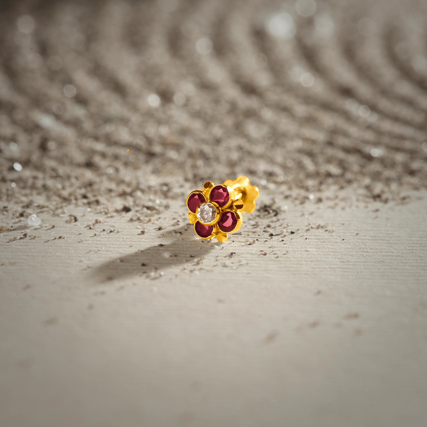 An 18K gold nose pin shaped like a flower with ruby-red petals and a clear crystal center, photographed on a sandy textured surface 
