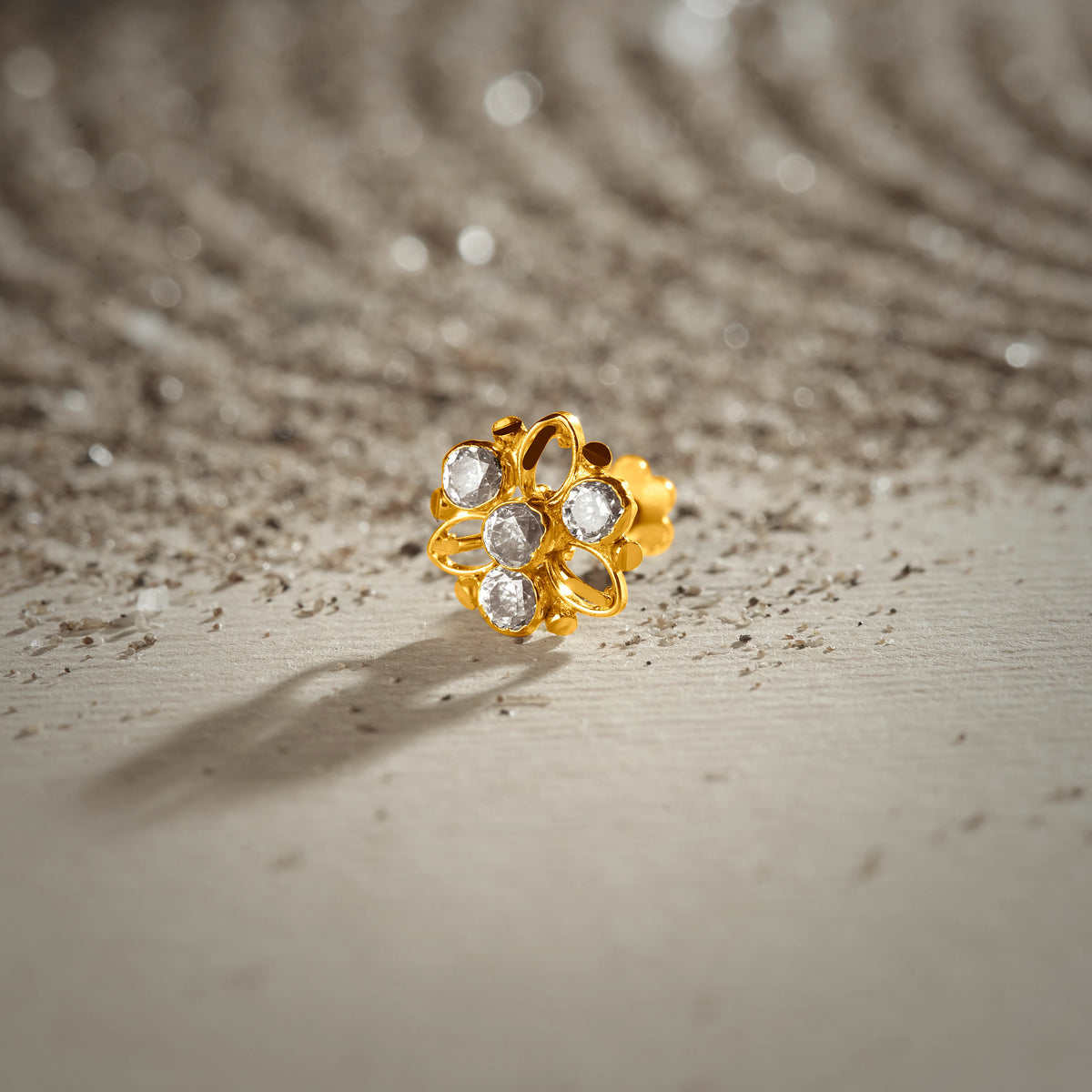 An 18K gold nose pin shaped like a flower with open gold petal outlines, set with clear crystal stones, photographed on a light textured surface