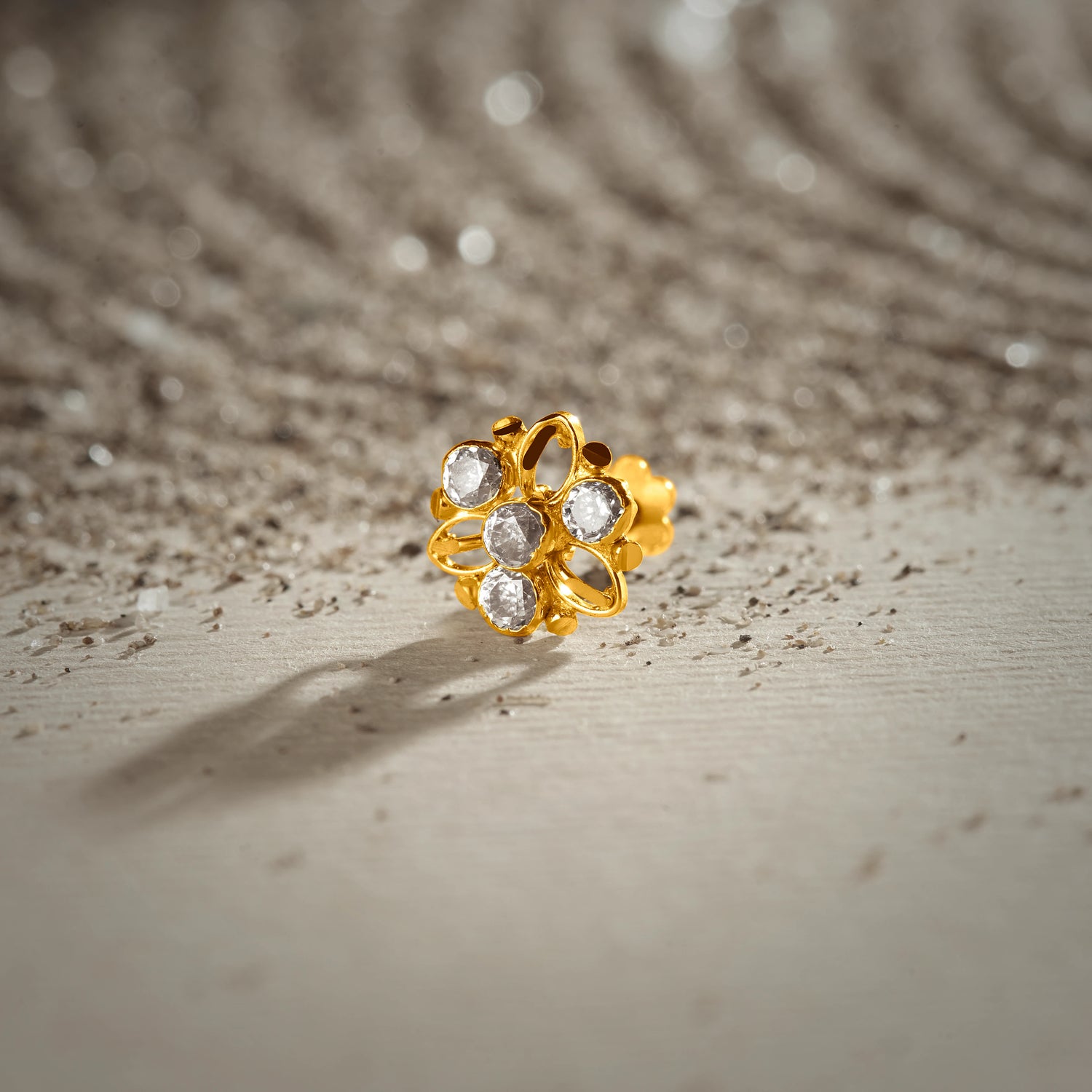 An 18K gold nose pin shaped like a flower with open gold petal outlines, set with clear crystal stones, photographed on a light textured surface