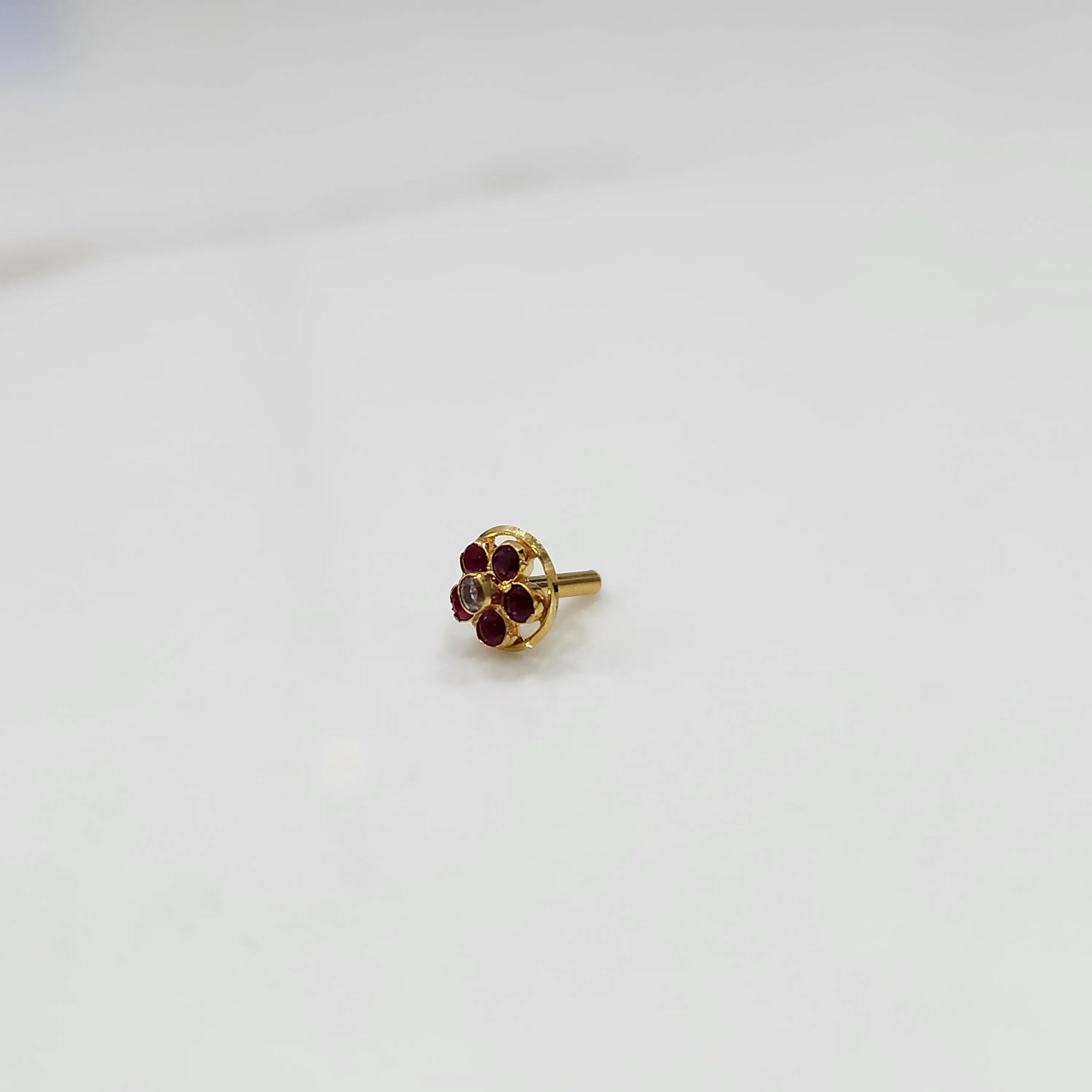 Product shot of a round gold nose stud with ruby red gemstone flower cluster