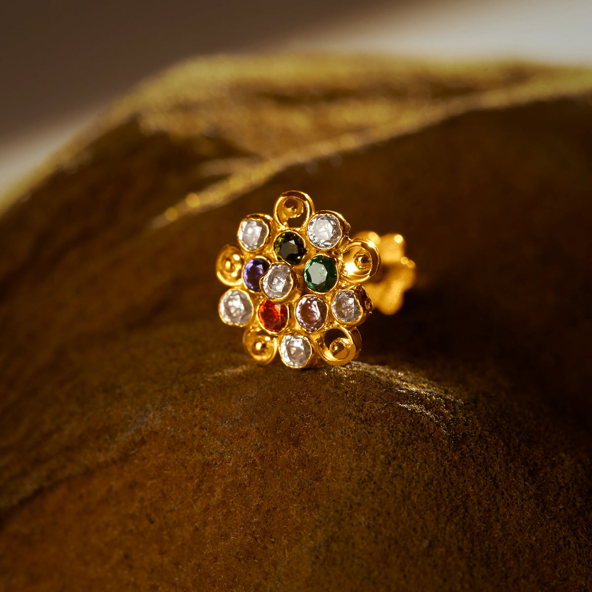 An 18K gold nose pin shaped like a flower with swirling loop edges, set with multicolored stones in green, purple, red, and clear, photographed on a dark textured fabric background