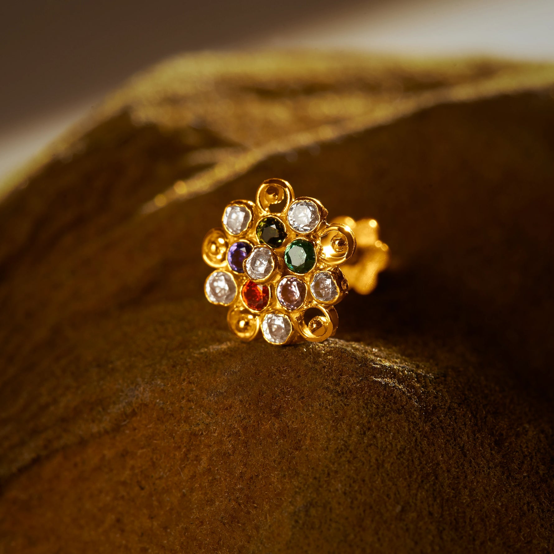 An 18K gold nose pin shaped like a flower with swirling loop edges, set with multicolored stones in green, purple, red, and clear, photographed on a dark textured fabric background