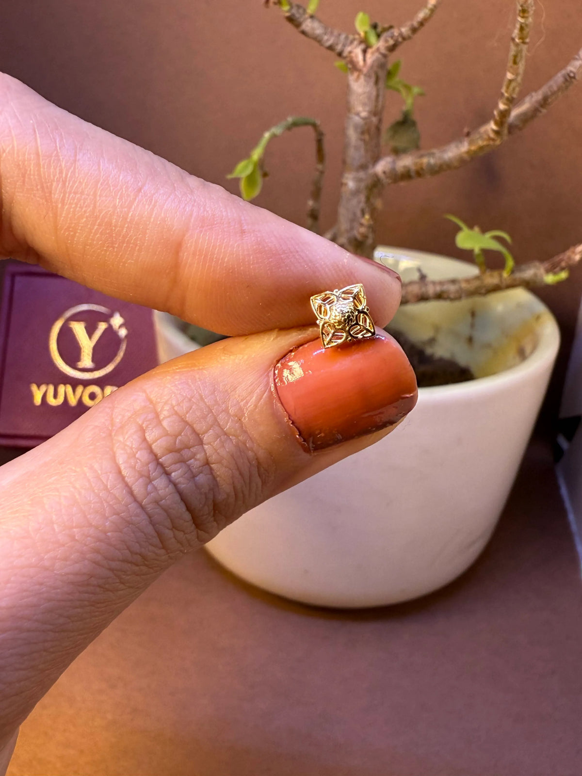 Square-shaped gold nose pin with filigree cutout design, held between fingertips