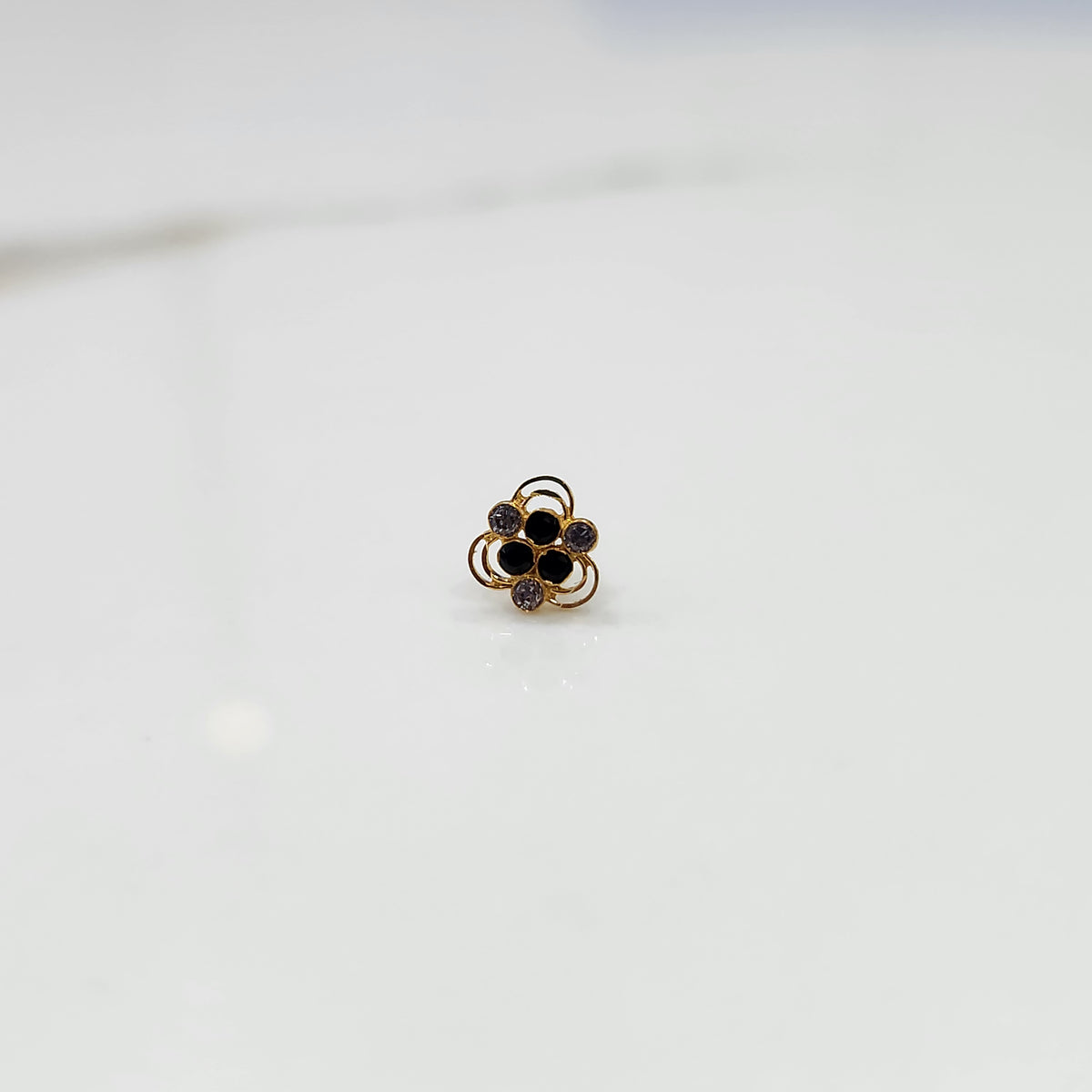 An 18K gold nose pin shaped like a flower, set with black and lavender-grey stones in open gold loops, close-up view