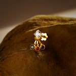 An 18K gold nose pin with a diamond-shaped clear crystal top and three hanging colored stones in green, red, and blue, photographed on a dark textured fabric background