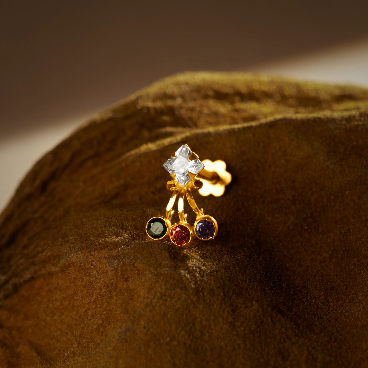 An 18K gold nose pin with a diamond-shaped clear crystal top and three hanging colored stones in green, red, and blue, photographed on a dark textured fabric background
