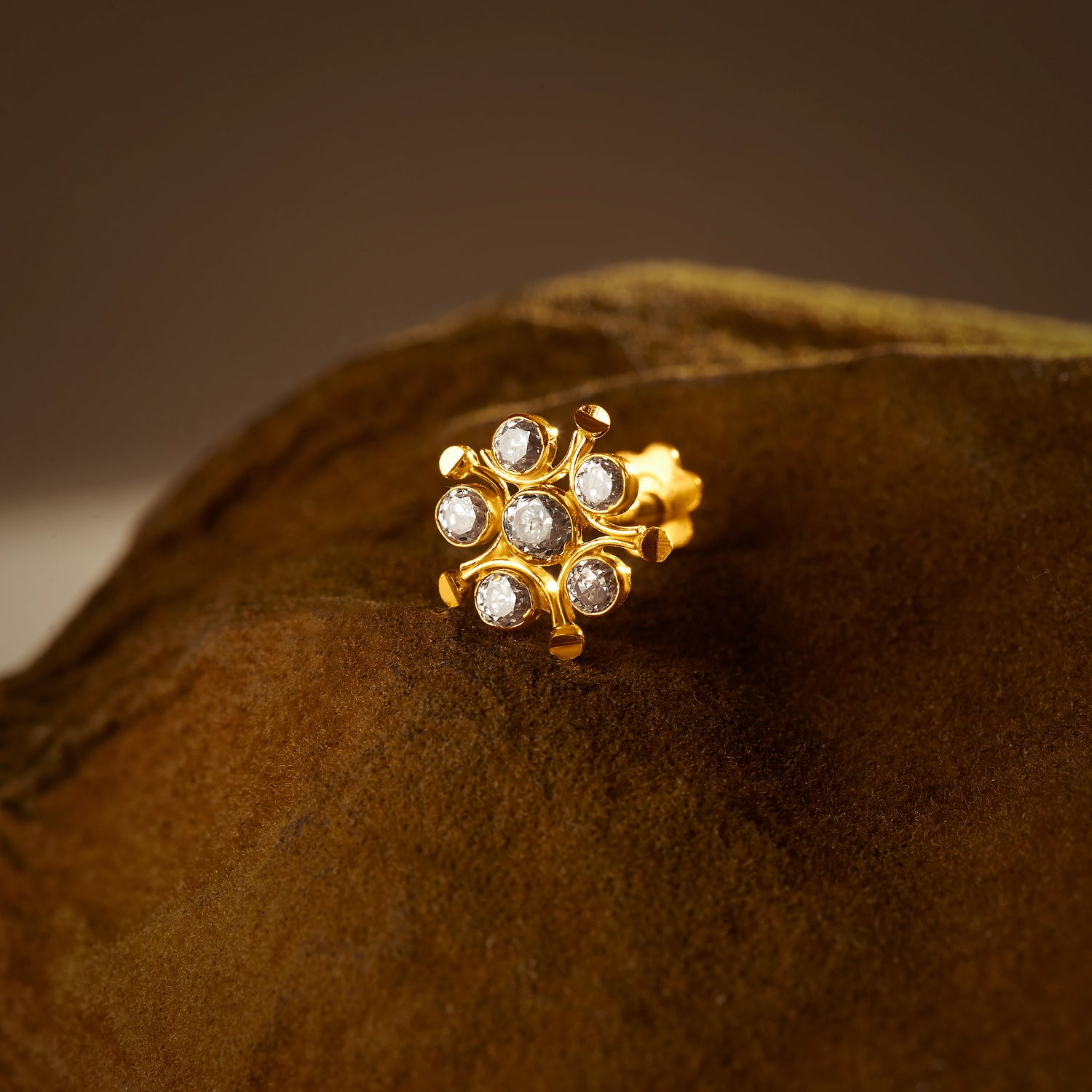 An 18K gold nose pin shaped like a sunburst, set with clear crystal stones, photographed on a dark textured fabric background