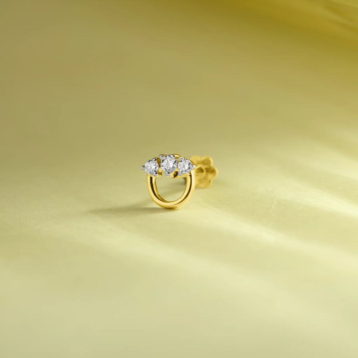 Round gold nose pin with three sparkling white stones on a soft pastel green background.