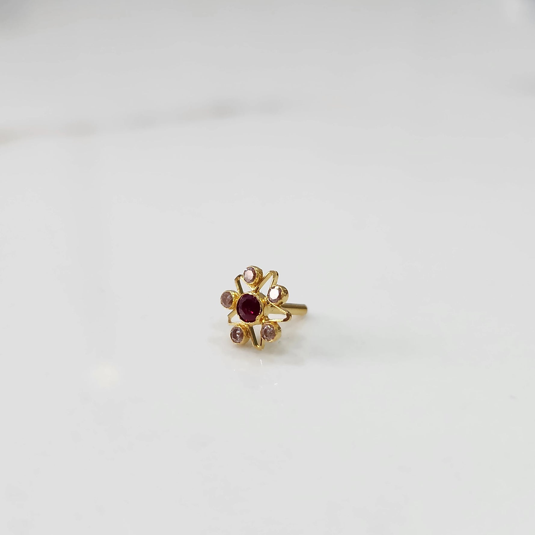 An 18K gold nose pin shaped like a star with a ruby-red center stone and light pink accent stones, photographed on a plain white background