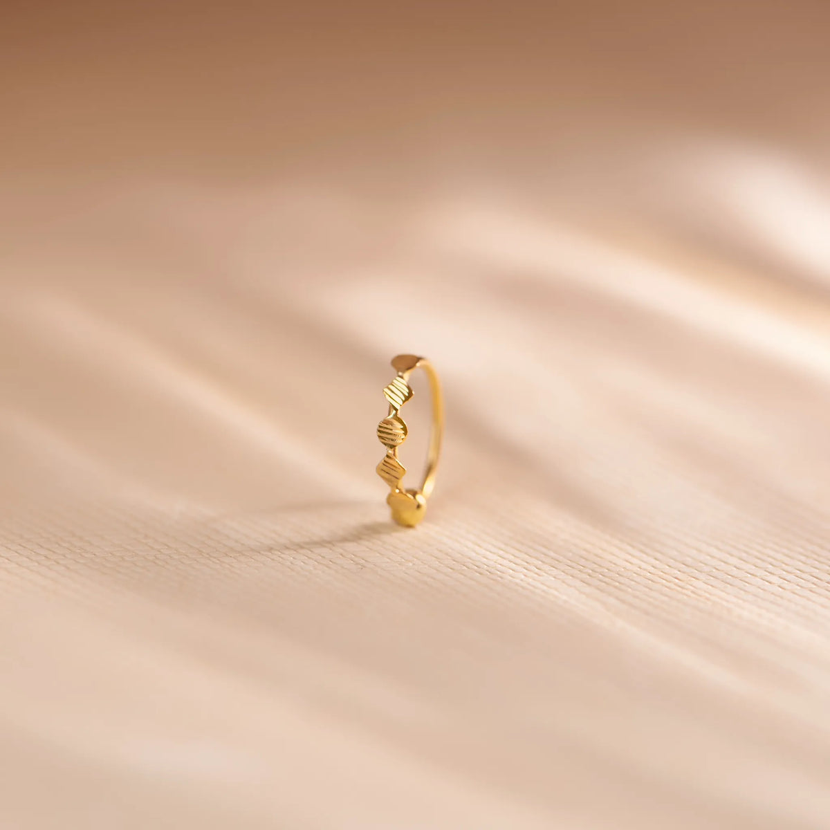 Close up of an 18K gold nose ring with sculpted wavy texture standing upright on a soft beige fabric background with gentle light streaks