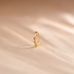 Close up of an 18K gold nose ring with sculpted wavy texture standing upright on a soft beige fabric background with gentle light streaks
