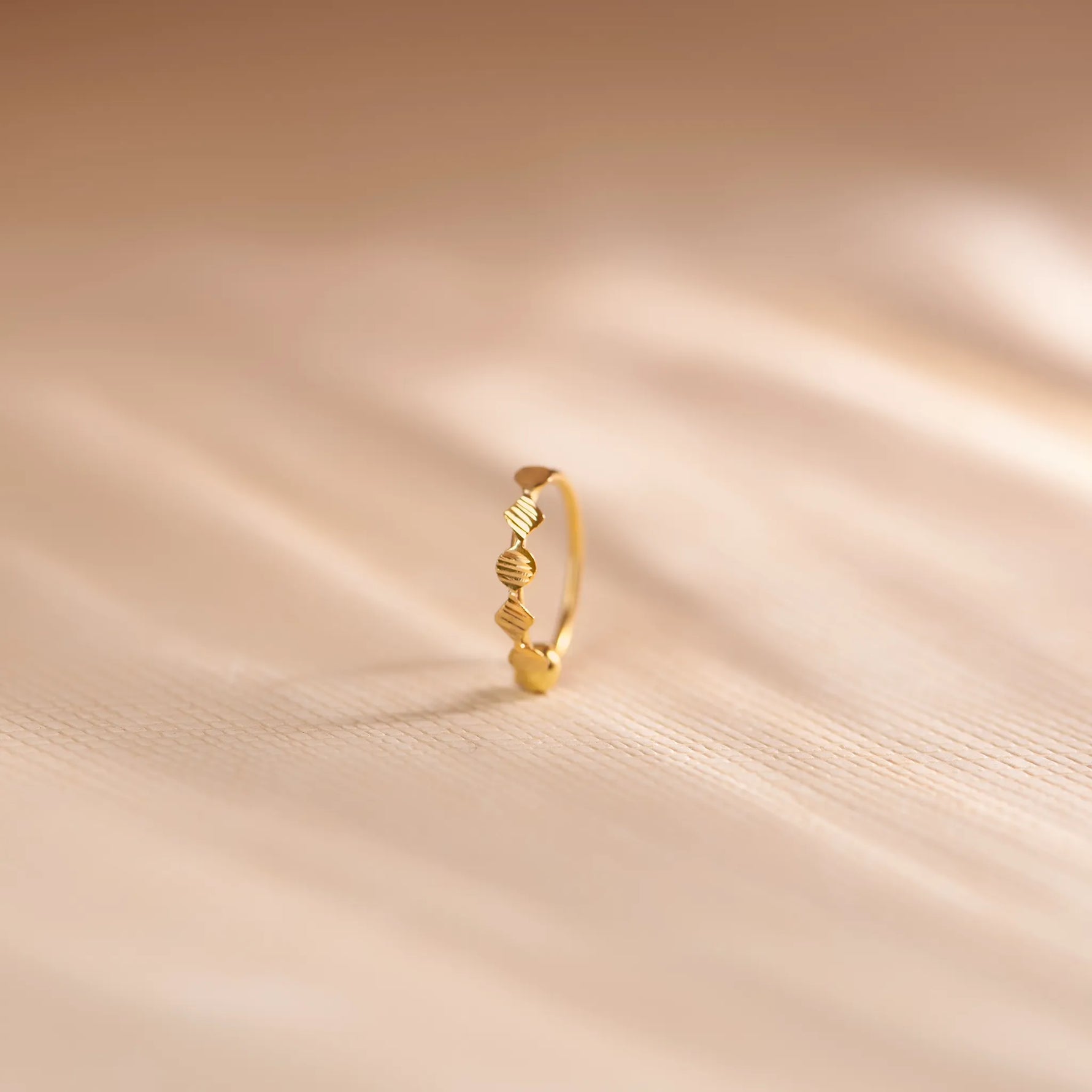 Close up of an 18K gold nose ring with sculpted wavy texture standing upright on a soft beige fabric background with gentle light streaks