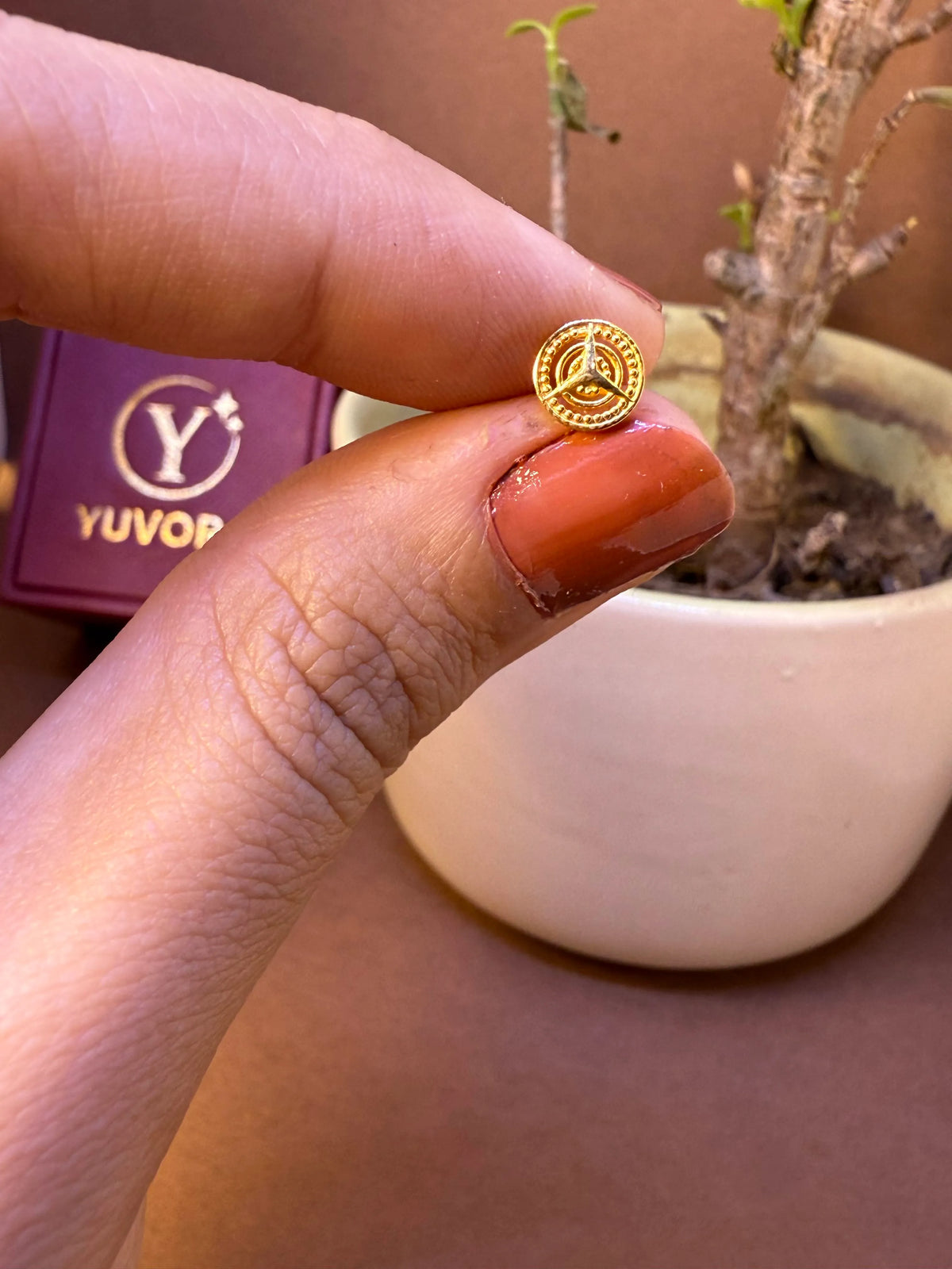Round gold nose pin with spoked wheel engraving, held between fingertips
