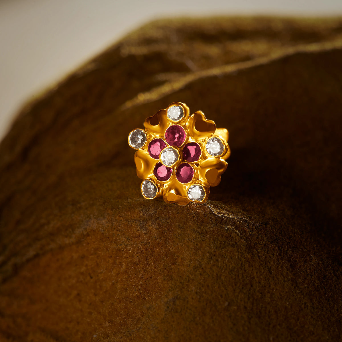 An 18K gold nose pin shaped like a dense flower cluster with small heart-shaped gold accents, set with alternating ruby-red and clear crystal stones, photographed on a dark textured fabric background