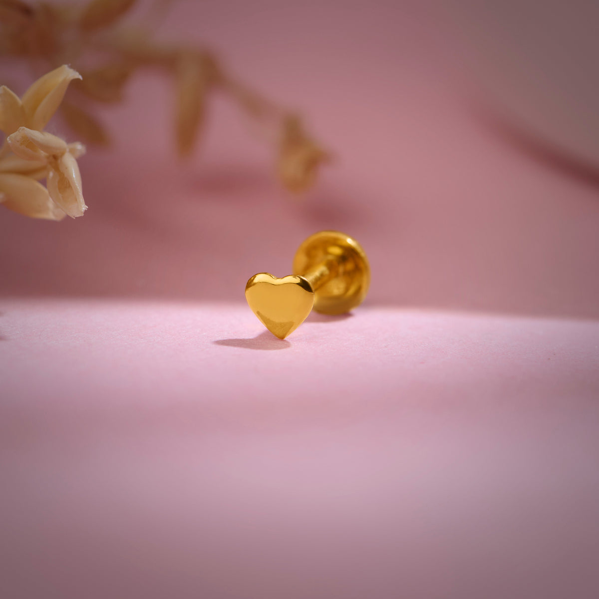 Close-up of heart-shaped 18K nose pin gold placed on a soft pink surface highlighting its smooth polished finish