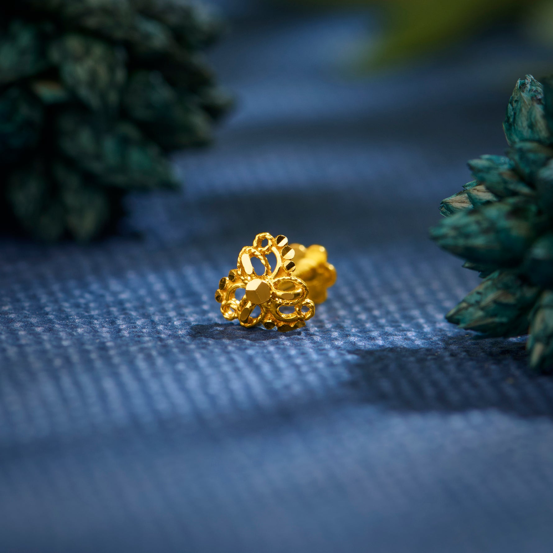 Close up of an intricate floral gold nose pin placed on a textured blue surface with greenery in the background highlighting its ornate design